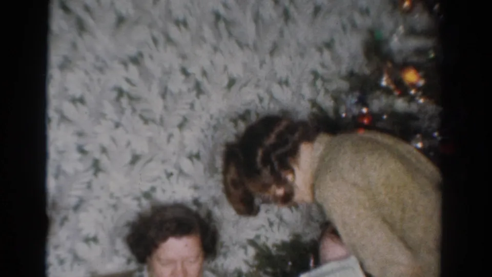 A younger woman bends over to help an older woman near the decorated tree in this 1950s Toledo living room scene.