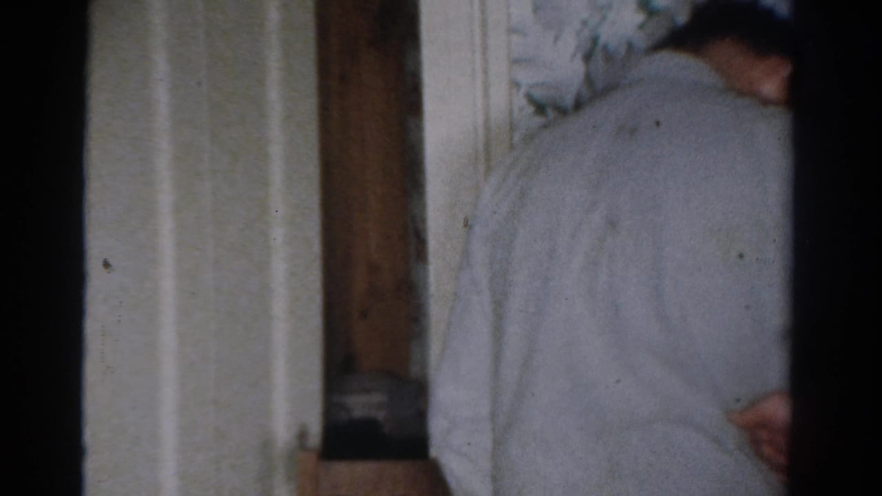 In a 1954 Toledo home interior, one adult turns away near floral wallpaper while a young man sits partly visible by a curtained window.