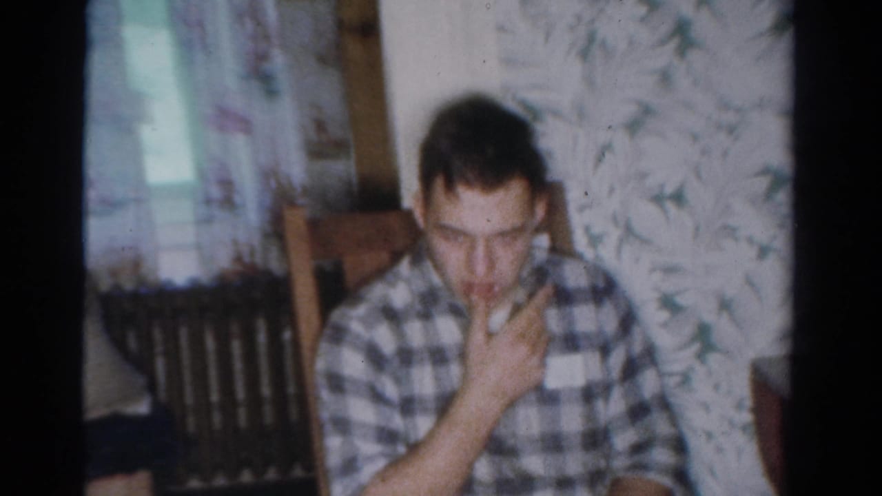 A young man in a plaid shirt sits in a wooden chair inside a 1954 Toledo home, holding his hand near his mouth as if eating or drinking.