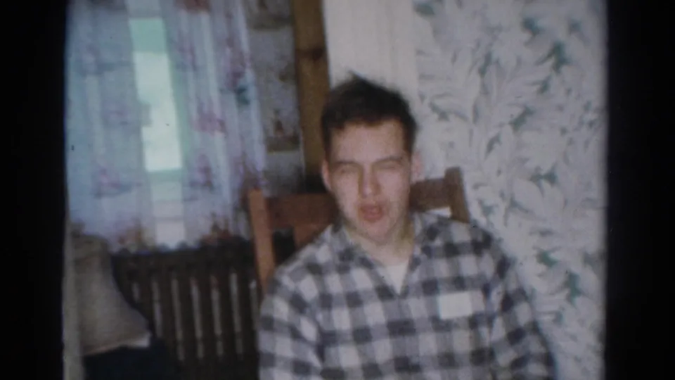 The young man faces toward the camera from a wooden chair in a Toledo home, with floral wallpaper, curtain, and radiator behind him.