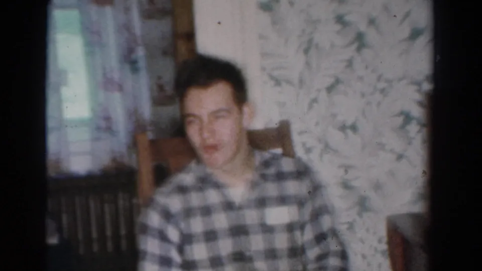 The young man sits back in the chair inside the 1954 Toledo home, appearing relaxed with a slight smile amid floral wallpaper and window light.