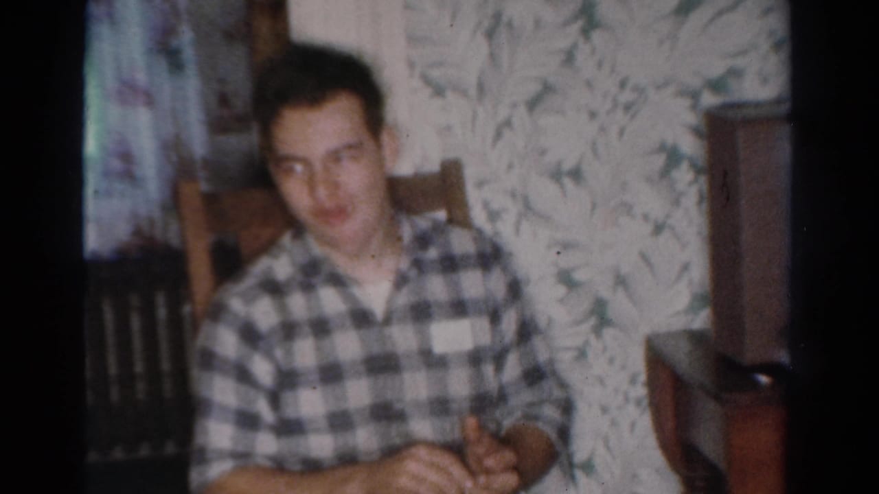 Young man in a plaid shirt sits in a Toledo, Ohio home interior with floral wallpaper, seen in 1954 8mm home-movie footage.