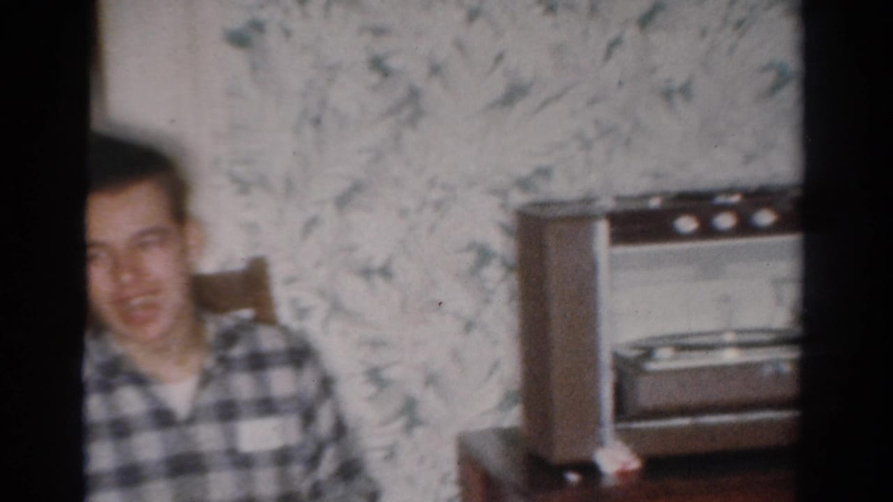 The seated young man appears beside floral wallpaper and a mid-century radio or record-player console in the Toledo home interior.