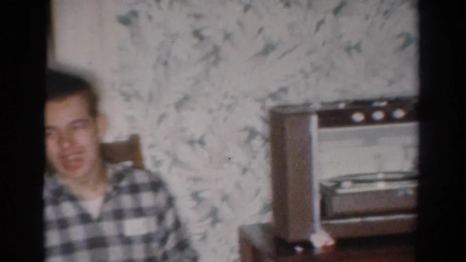 The seated young man appears beside floral wallpaper and a mid-century radio or record-player console in the Toledo home interior.