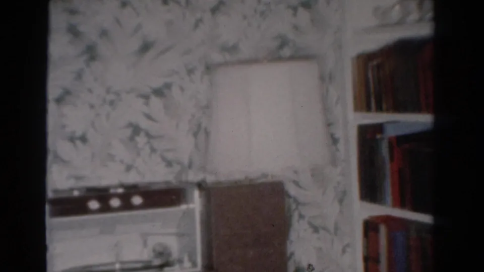 A Toledo home interior shows a radio or record-player console, table lamp, floral wallpaper, and bookshelf in 1954 8mm footage.