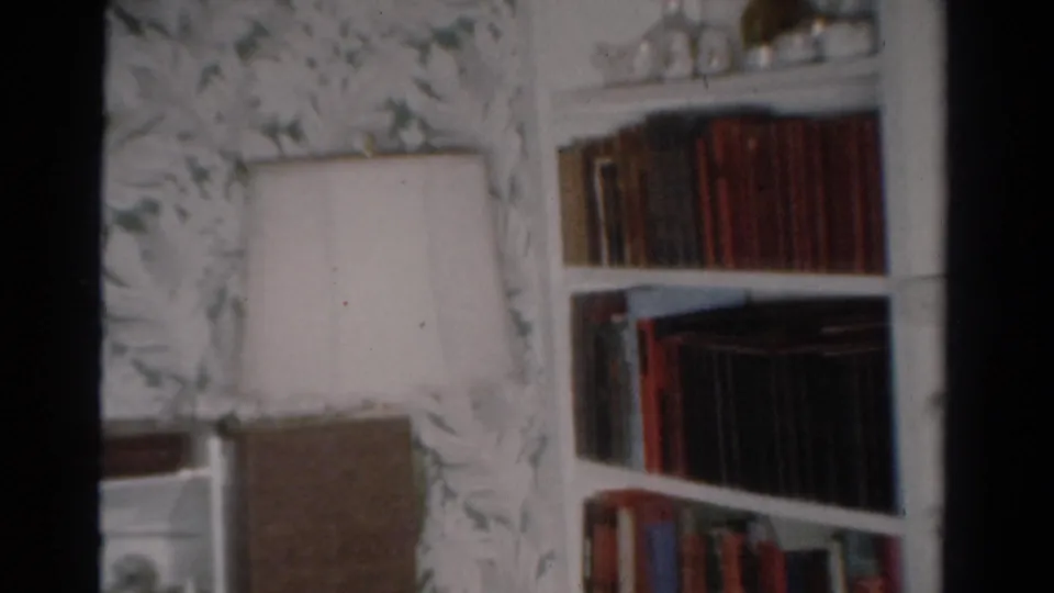 1954 Toledo home-movie frame showing a bookshelf, white lampshade, and floral wallpaper in a private living room.