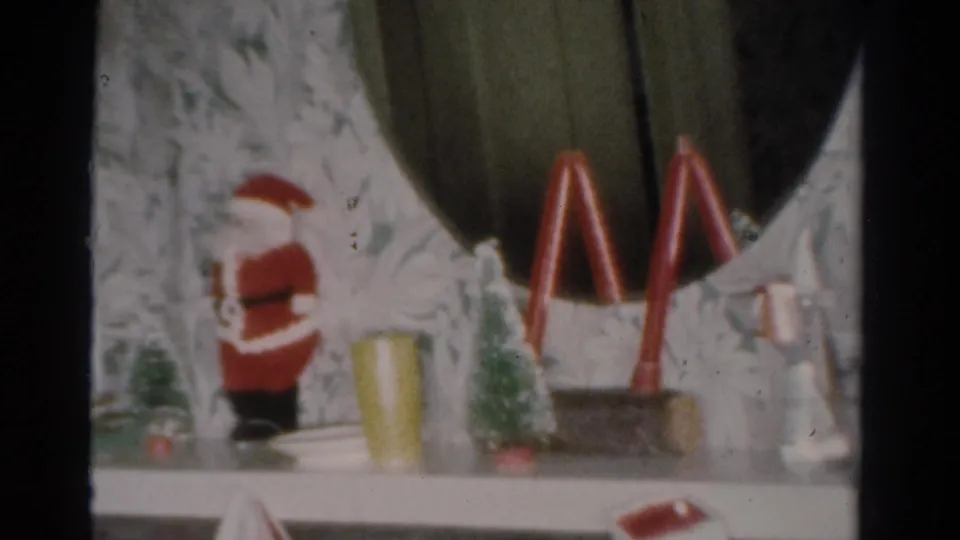 1954 Toledo 8mm view of Christmas mantel decorations, including Santa, small trees, red arches, a yellow cup, and stockings.
