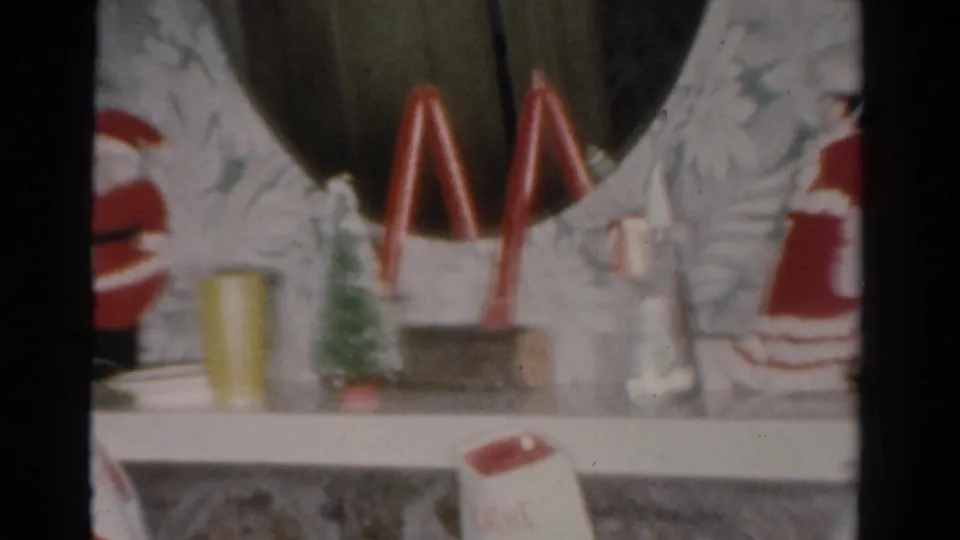 1954 Toledo home-movie frame showing a mantel display with red arches, miniature trees, a partial Santa figure, white figurine, and stockings.