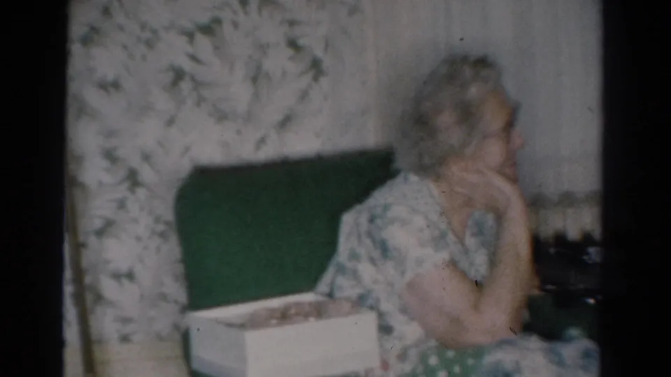 1954 Toledo home-movie frame of an older woman in a floral dress seated on a green couch inside a decorated private home.