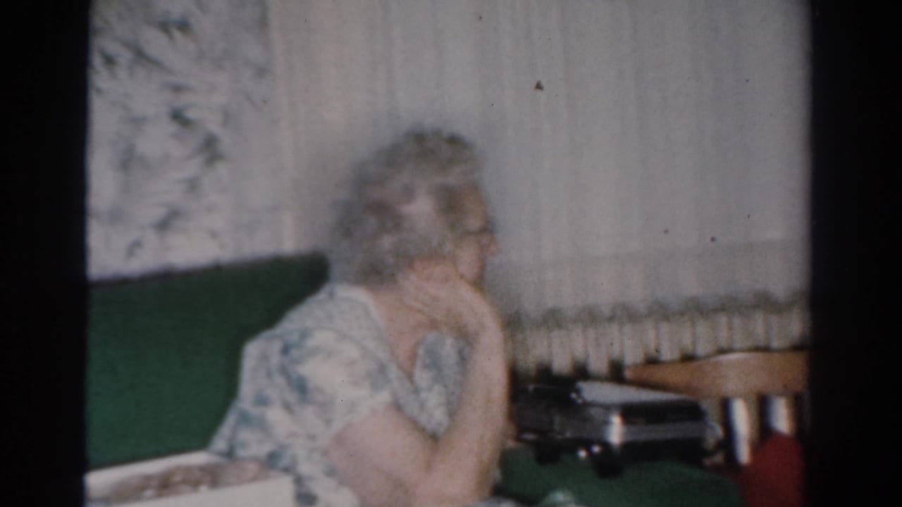 Woman in glasses sits in a living room with her hand near her chin.