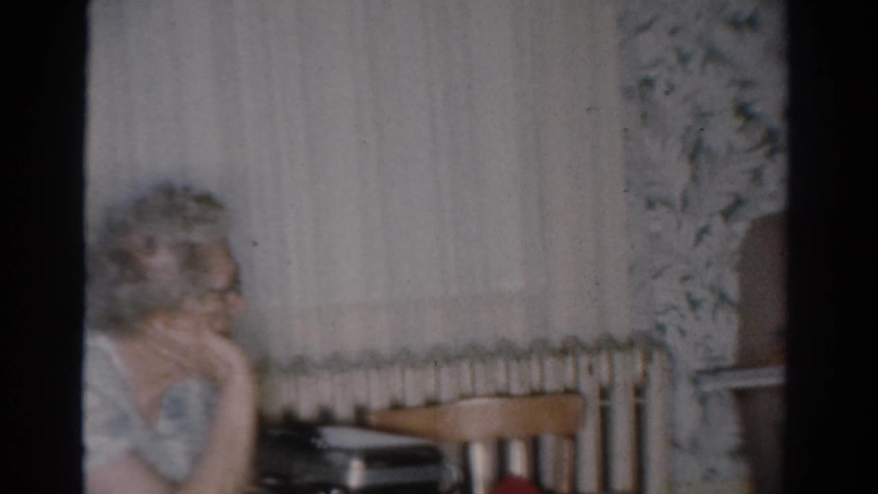 The woman remains seated in the Toledo living room, looking sideways past a radiator, curtains, and wooden chairs.