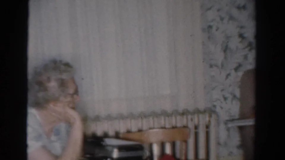 The woman remains seated in the Toledo living room, looking sideways past a radiator, curtains, and wooden chairs.