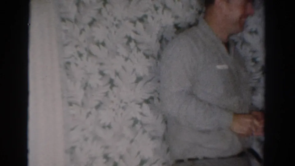 A man in a light-colored sweater stands near floral wallpaper inside the 1954 Toledo living room.