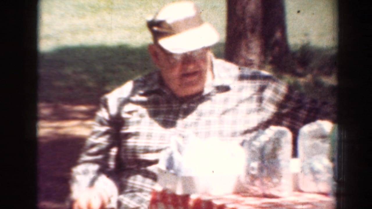 Elderly man in a plaid shirt and light cap sits at an outdoor picnic table in 1954 Toledo home-movie footage.