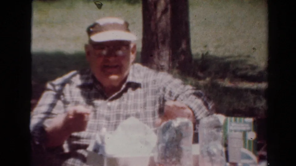 The elderly man faces the camera while holding food or a skewer at the picnic table in the 1954 home movie.