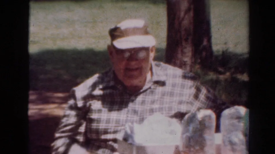 The man appears to smile toward the camera at the picnic table, with trees and picnic items around him.