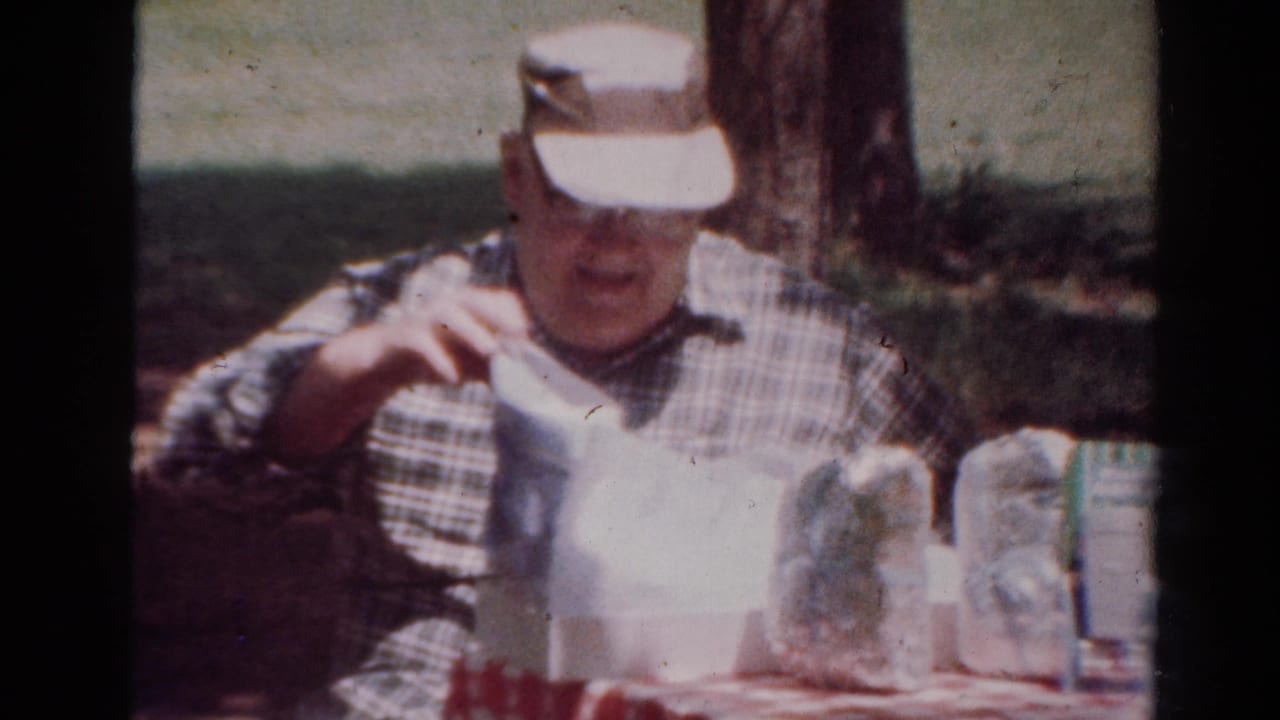 In this 1954 Toledo home-movie frame, a man in a plaid shirt and white visor sits outdoors at a table with food packages and a checkered cloth.