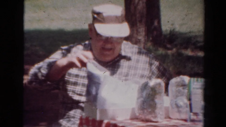 In this 1954 Toledo home-movie frame, a man in a plaid shirt and white visor sits outdoors at a table with food packages and a checkered cloth.