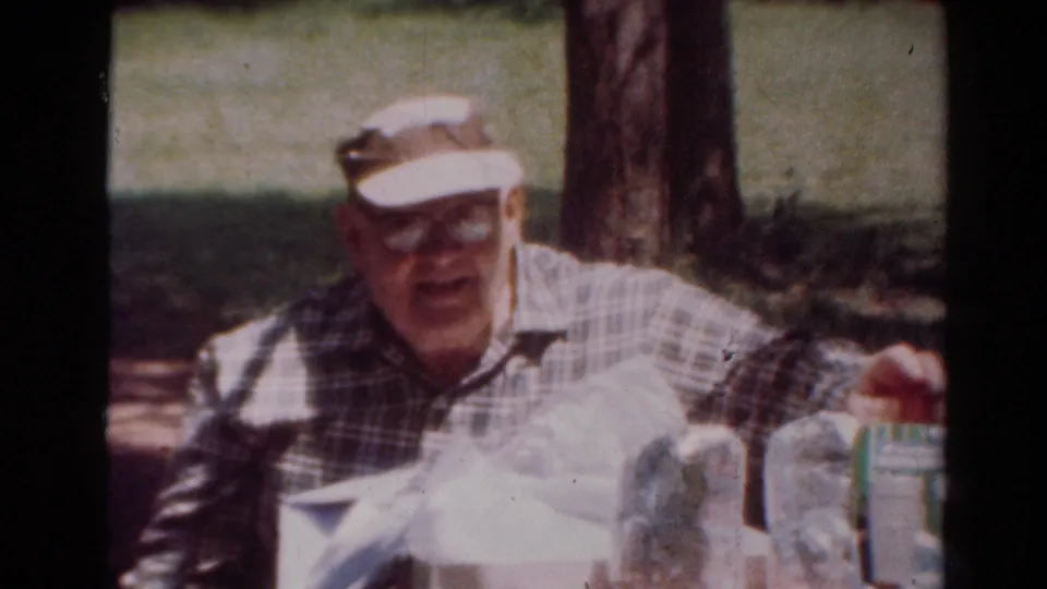 The man leans forward toward the camera at an outdoor table with a carton, bag, and packaged items in this 1954 Toledo home-movie frame.