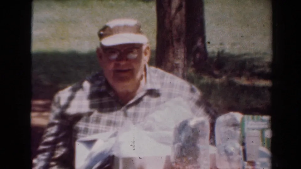The man faces the camera from behind the checkered outdoor table, with packages and a light-colored bag visible in the 1954 Toledo footage.