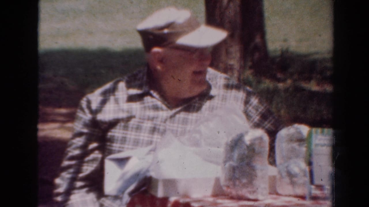 In 1954 Toledo home-movie footage, an older man in a plaid shirt and cap sits at a shaded picnic table with drinks and tableware.