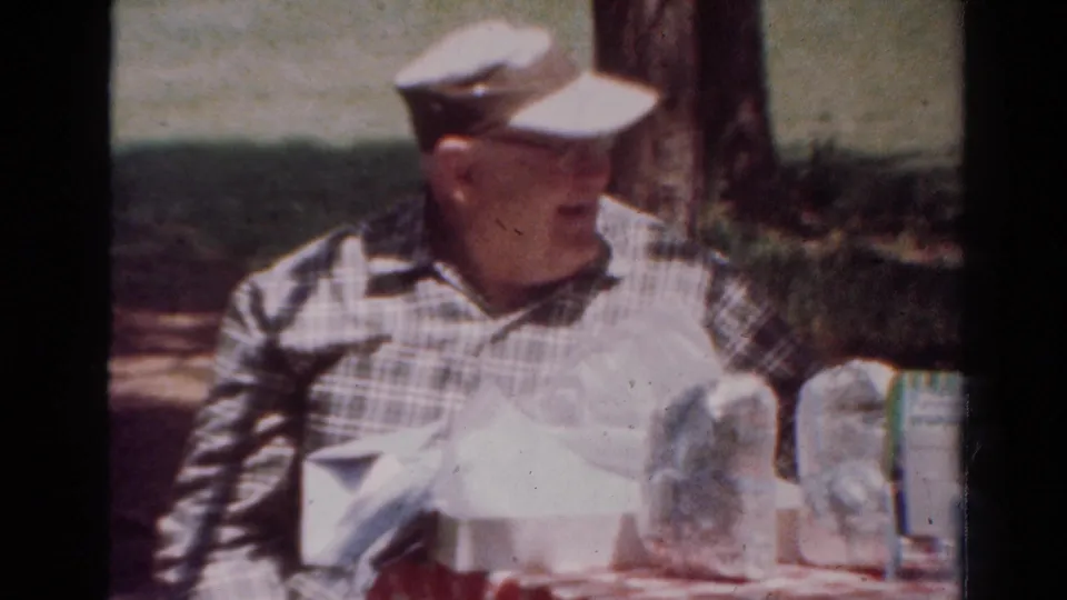 In 1954 Toledo home-movie footage, an older man in a plaid shirt and cap sits at a shaded picnic table with drinks and tableware.