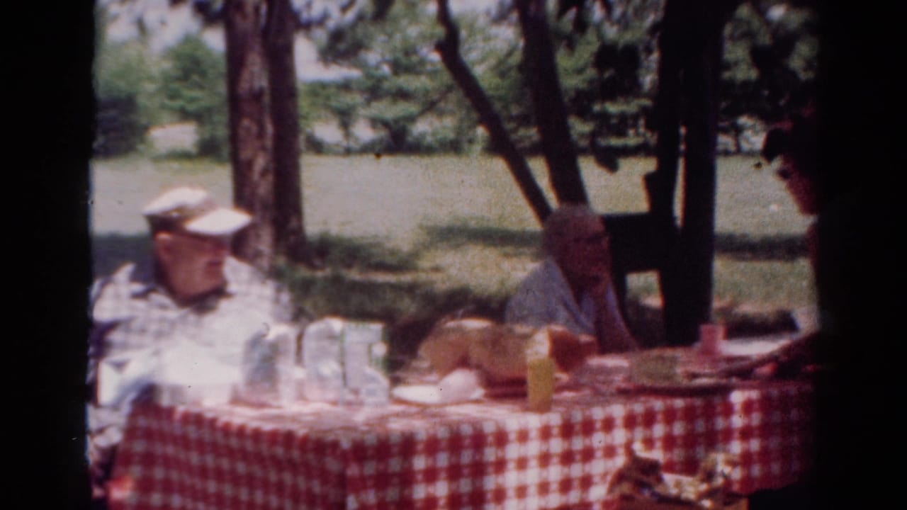 1954 Toledo home-movie frame of an older man in a white cap seated beside a checked picnic table under trees.