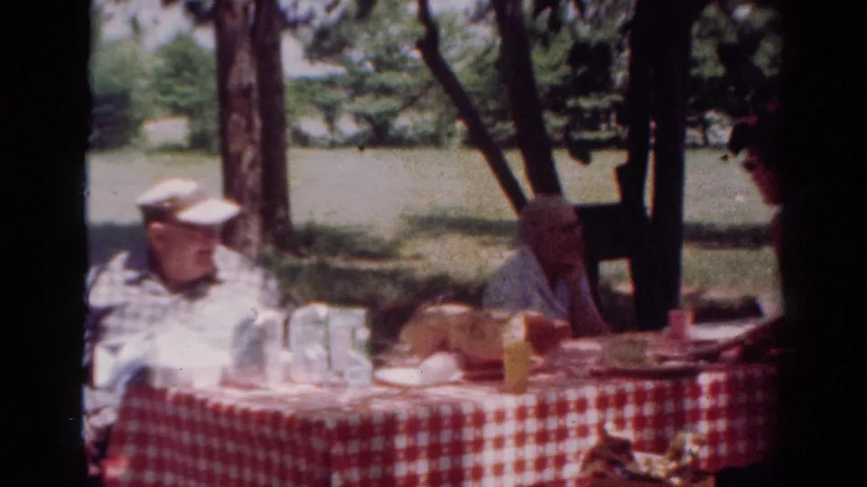 1954 Toledo home-movie frame of an older man in a white cap seated beside a checked picnic table under trees.