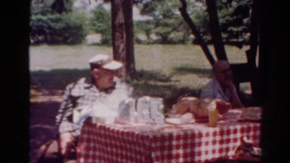 1954 Toledo outdoor picnic scene with an older man in a cap and another adult seated across a checked table.