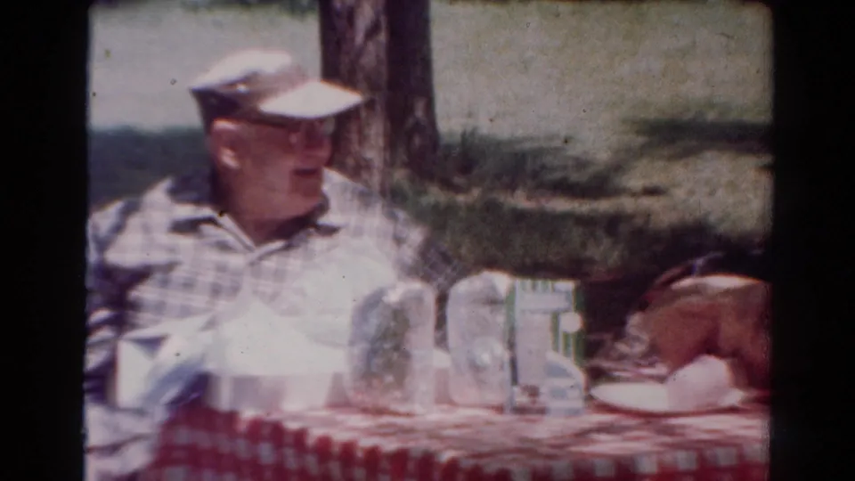 Closer 1954 Toledo home-movie view of an older man in plaid at a picnic table with jars, cups, and food.
