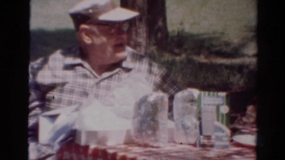 1954 Toledo frame of an older man in a plaid shirt and cap looking to the side during an outdoor picnic.