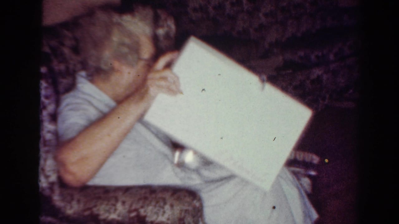 Elderly woman with glasses sits on a patterned couch holding a white gift box.