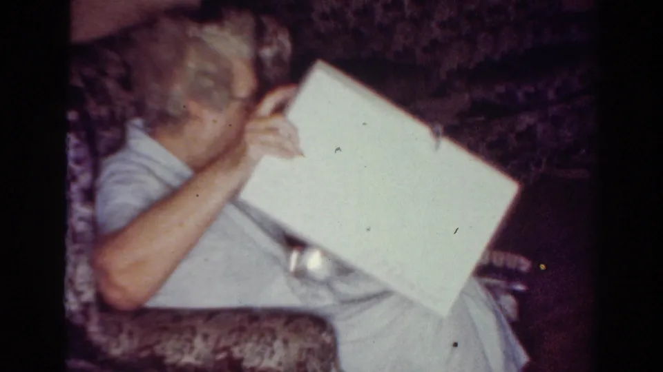 Elderly woman with glasses in a Toledo home interior in 1954 sits on a patterned couch and lifts a white gift box.