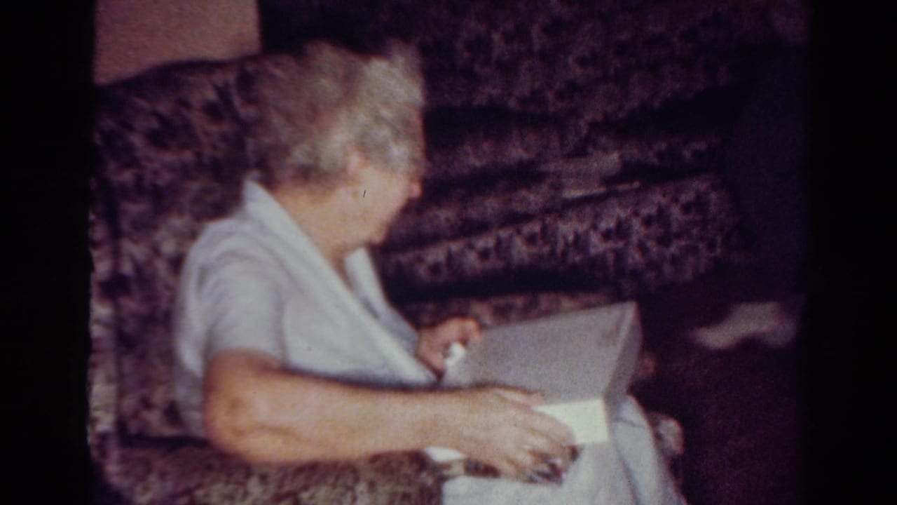 Elderly woman turns toward a white box while seated on a patterned couch.