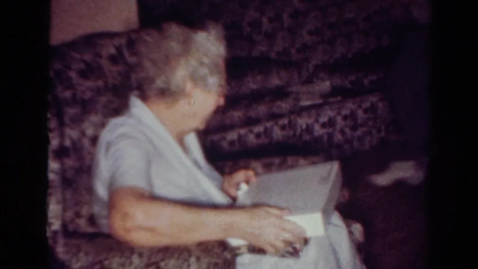 In the 1954 Toledo home-movie scene, the woman looks down toward a white box on her lap while seated on the couch.