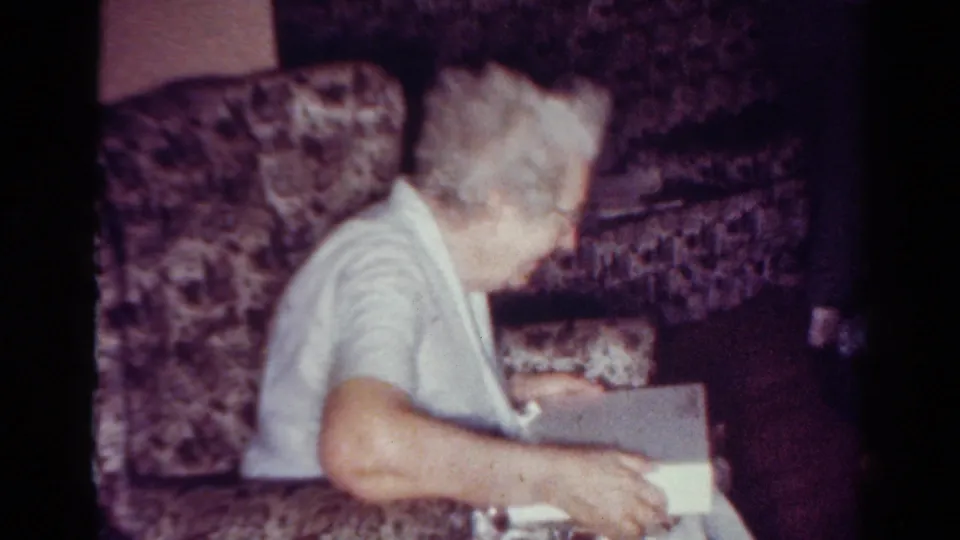 Side view of the elderly woman in the Toledo interior holding the white gift box while seated against patterned upholstery.
