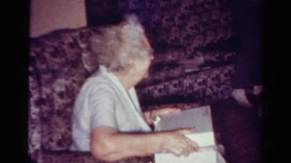 The woman remains seated in profile on the patterned couch with the white gift box open or partly open near her lap.