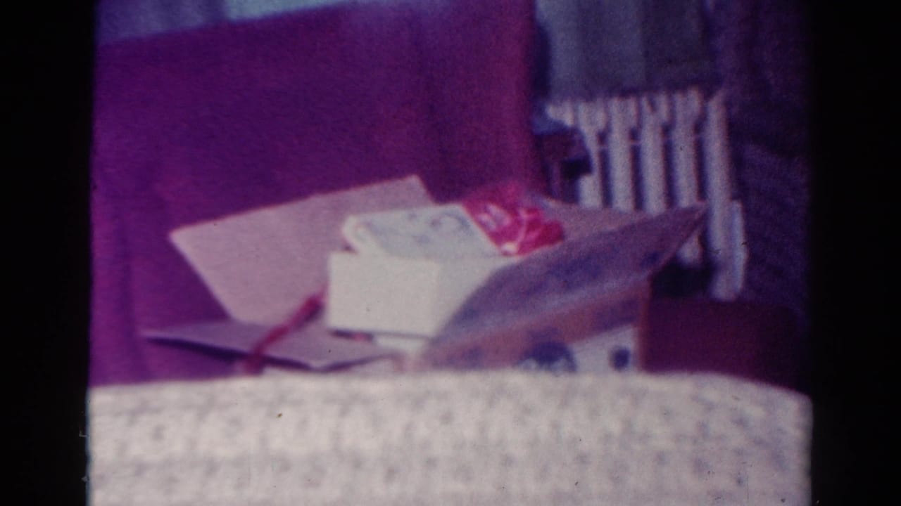 1954 Toledo home-movie frame showing a patterned couch, gift boxes, wrapping paper, and a wall radiator in a holiday living room.