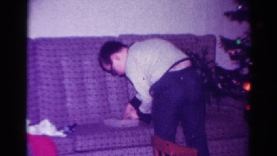 1954 Toledo home-movie frame showing a person leaning over a patterned couch beside a small lit Christmas tree.