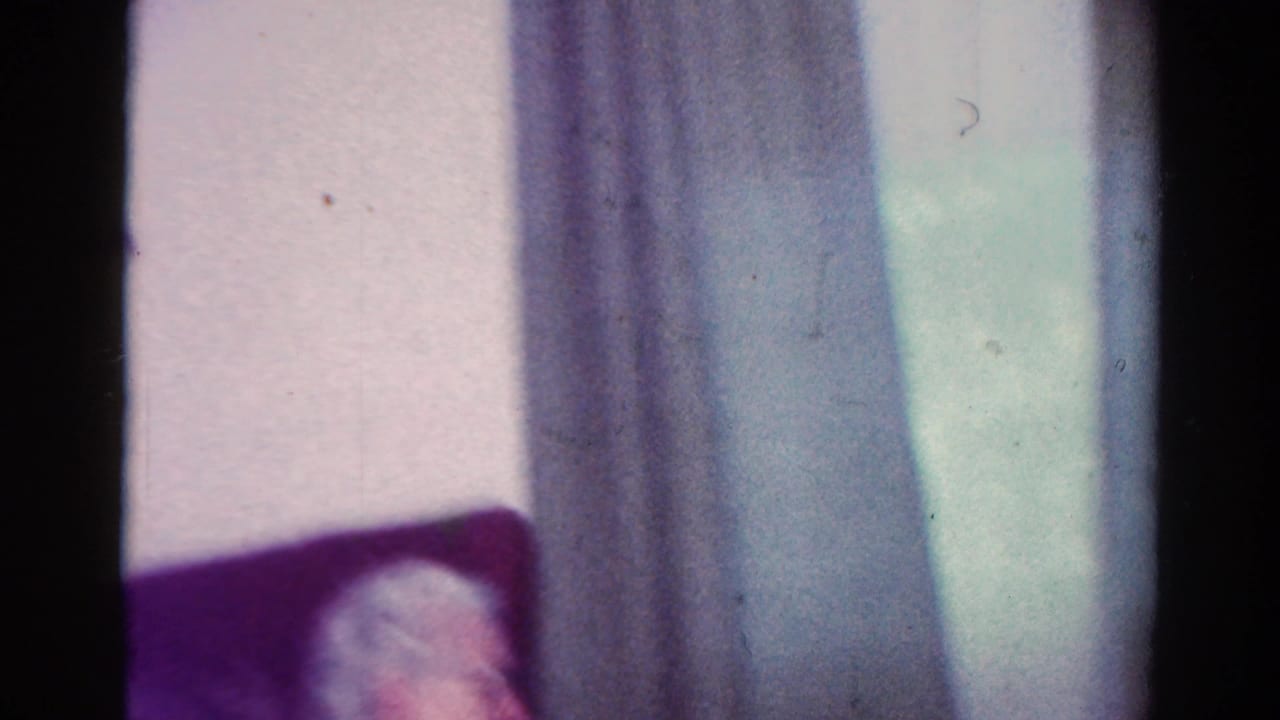 1954 Toledo home-movie frame showing an older man with glasses seated in a red upholstered chair near curtains.