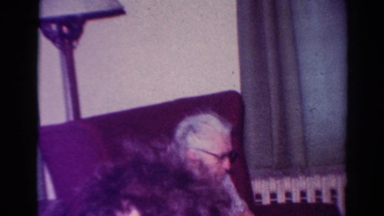 Older man in glasses seated in a red chair, partly obscured by a foreground head.