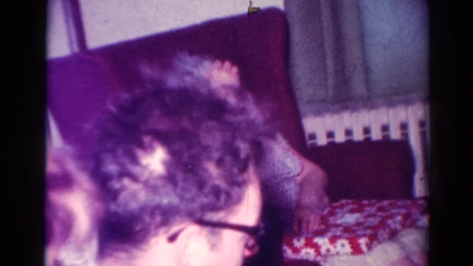 1954 Toledo home-movie frame showing a seated family member with glasses in the foreground and an older man on a red chair nearby.