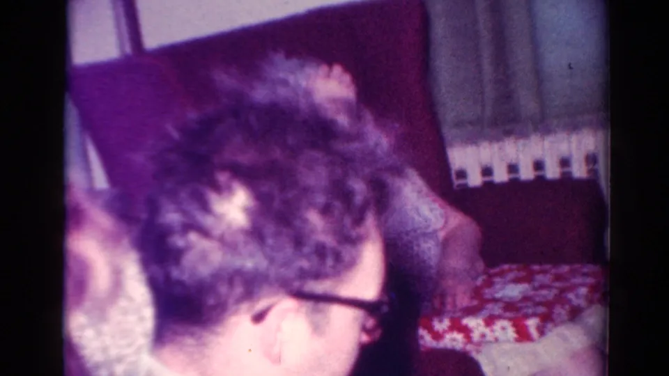 In a 1954 Toledo home-movie frame, a man with glasses sits close to a red-and-white patterned gift box in a living room.
