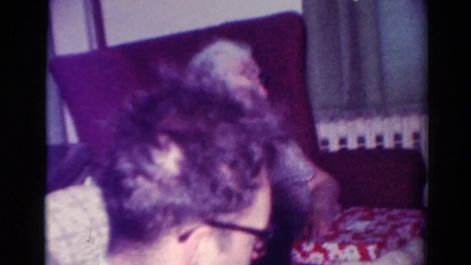 The frame shows the man bending forward in a Toledo living room while an older adult sits behind him near the radiator and curtains.