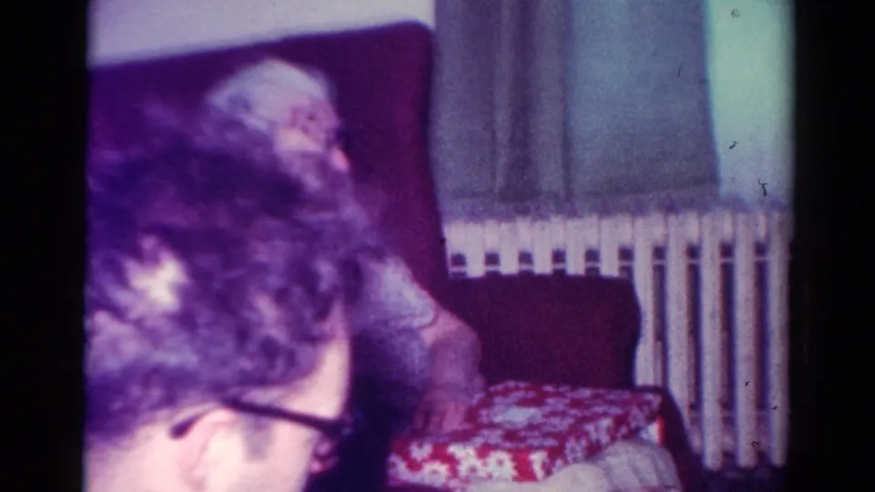 In the final frame, the man remains leaned forward while another seated adult and the red-and-white patterned box are visible in the living room.