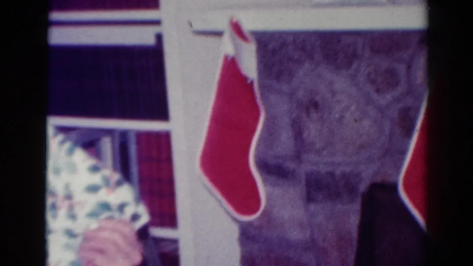 1954 Toledo home-movie frame showing red stockings by the fireplace with a holiday-wrapped gift partly visible at left.