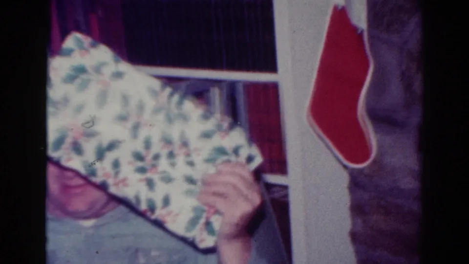 1954 Toledo home-movie frame showing a young person partly hidden by a wrapped gift beside a red Christmas stocking.