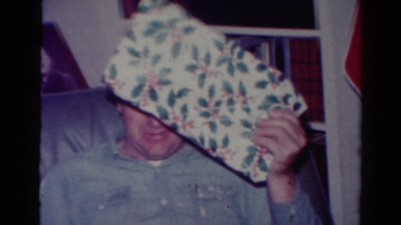 Person in a light sweater holds a holly-patterned wrapped gift near their face indoors.