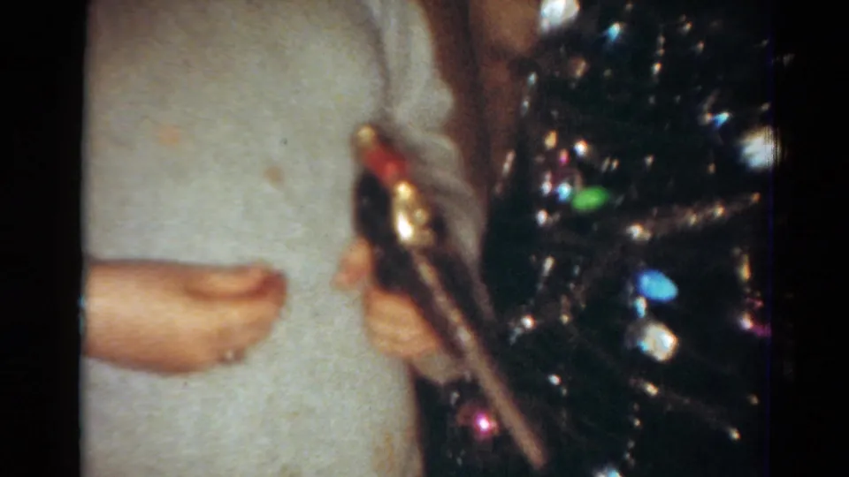 1954 Toledo home-movie frame showing cropped hands in a light sweater handling a toy gun near a shiny Christmas tree.