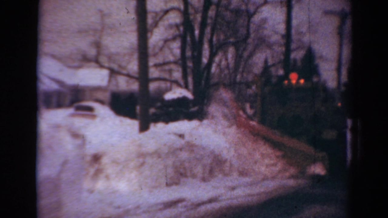 1954 Toledo frame with a snowplow truck moving along a residential road lined by snowbanks, trees, and houses.